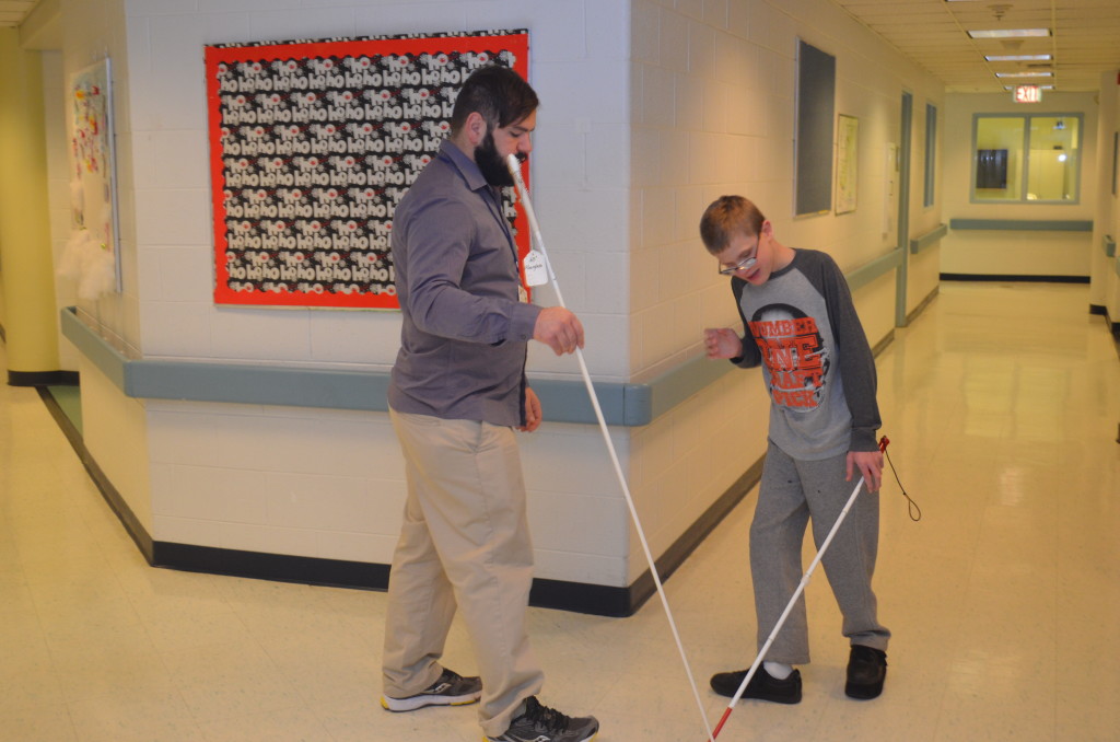 A mobility training instructor practices walking with a white cane to a young boy who is blind