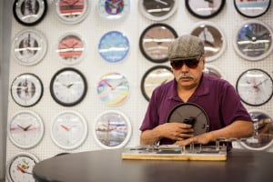 A man who is blind assembles a clock. In the background is a colorful display of fasion clocks.