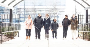 A group of six individuals walking under a glass archway as they enter The Chicago Lighthouse. All of the individuals have a disability, either blindness, deafness or cerebral palsy