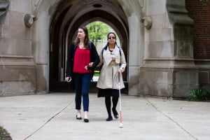 Two young women walk through a courtyard. One is blind and uses a white cane.