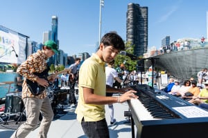 A teenage musician plays the keyboard with his band at an outdoor concert at Navy Pier. A crowd watches against a beautiful Chicago skyline backdrop