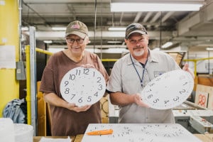 Two men working in the clock factory hold up clock faces before they are inserted into the frames