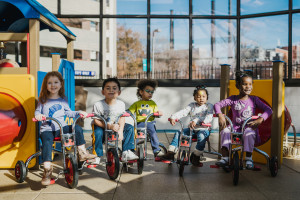 Five children on tricycles play in a sunny solarium. Two of the children have a significant visual impairment.