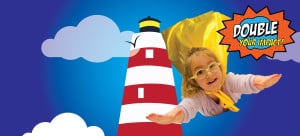 A preschool girl wearing glasses and a yellow superhero cape fly through a cloudy sky around a lighthouse. Double Your impact sign is beside her.