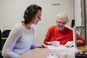 An occupational therapist demonstrates the benefit of wide lined paper, thick black markers and task lighting to an older client who is visually impaired