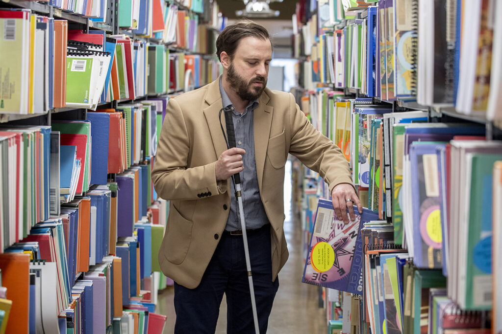 A man who is blind stands in an aisle of the Illinois Instructional Materials  Center and pulls out a large print book