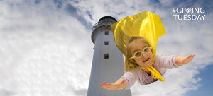 A preschool girl wearing glasses and a yellow superhero cape fly through a cloudy sky around a lighthouse