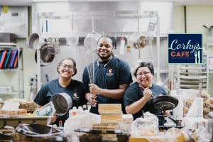 3 employees of Corky's Cafe pose by the counter with a pot, pan and oversized wisk