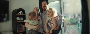 A person holds two young children inside a living room, all smiling and appearing joyful.