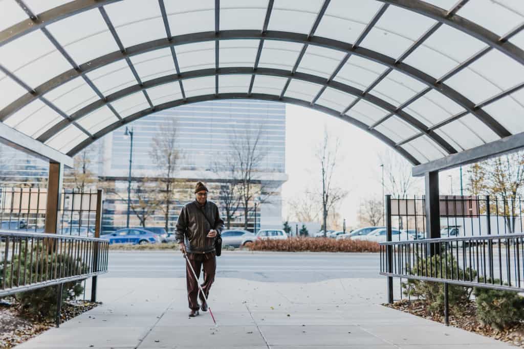 An individual using a white cane walks on the sidewalk. 