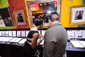 Two people browse silent auction bid sheets displayed on a table.