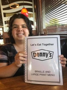 Sandy is shown smiling and holding up a menu Braille and Large Print Denny's Menu