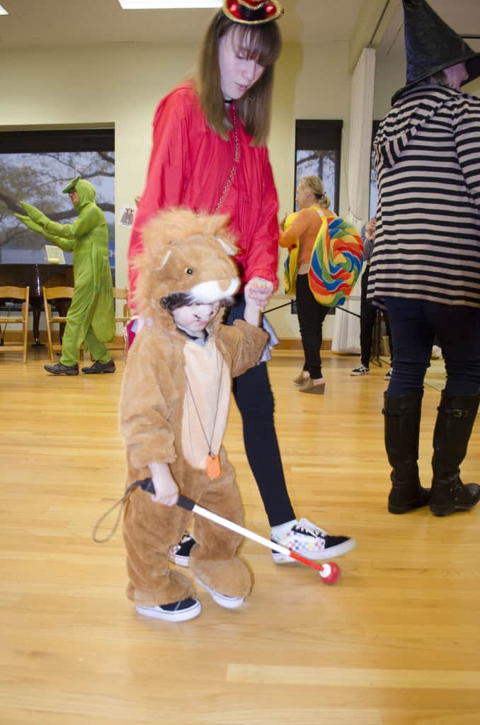 A young boy walks using a white cane while dressed like a lion. He holds hands with his older sister who is dressed like a ringmaster. 