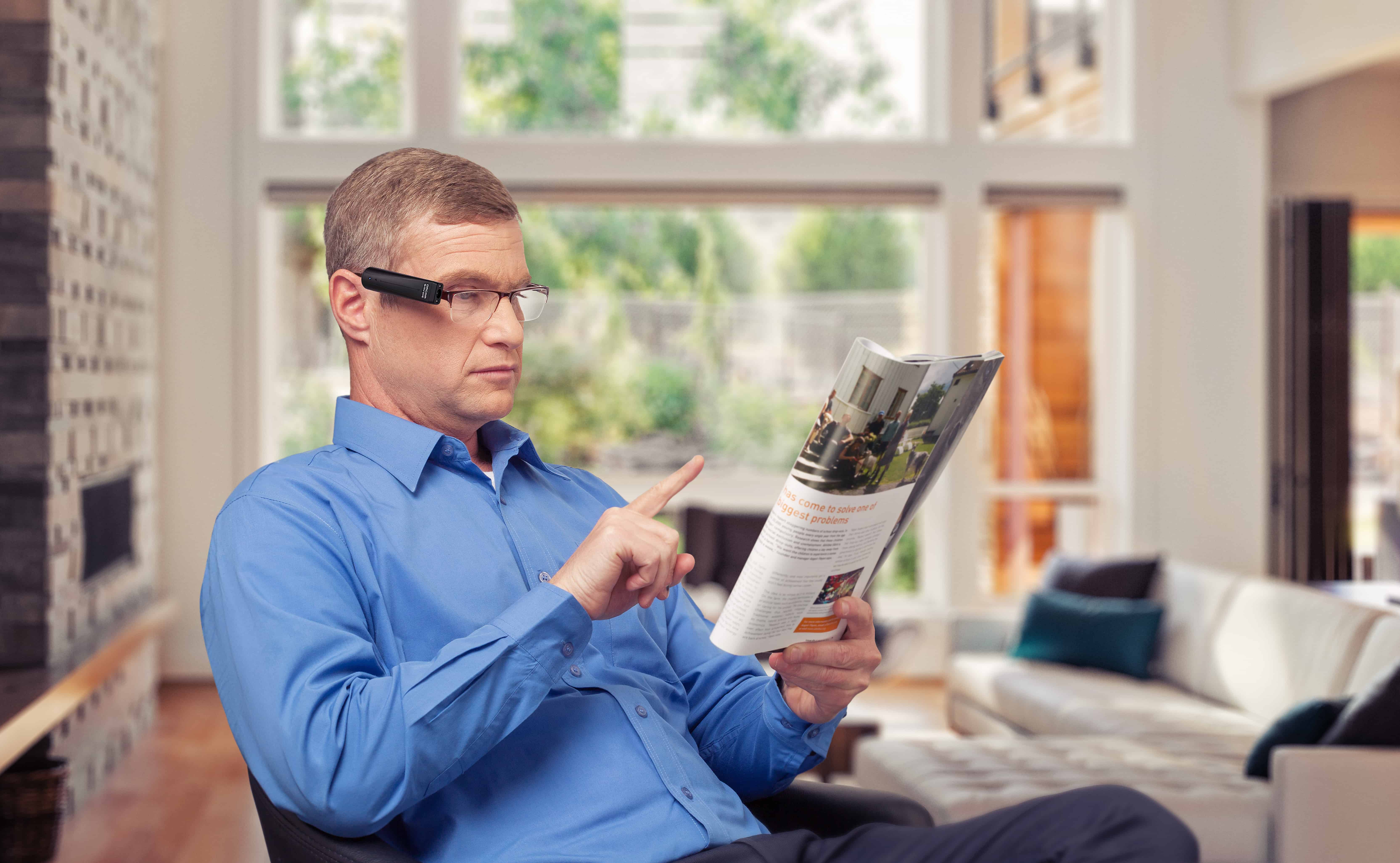 A man wearing OrCam's MyEye2 points to a position on a magazine to have it read aloud to an earpiece he is wearing.