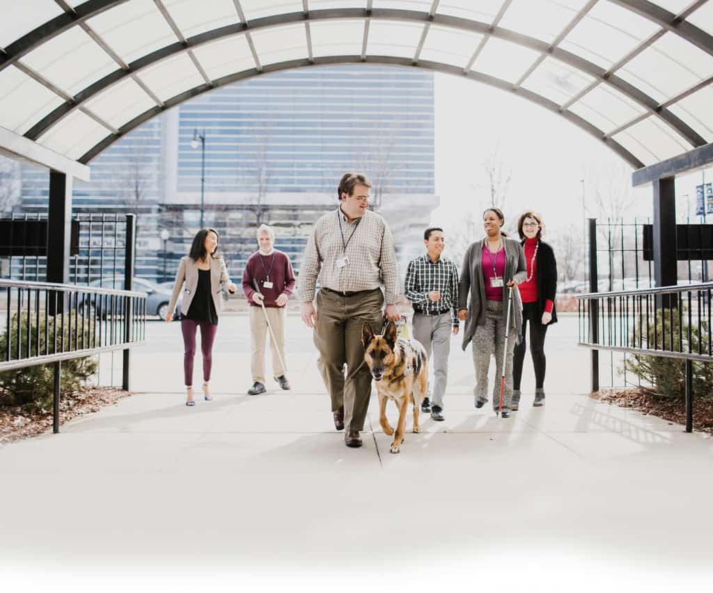 A group of six Chicago Lighthouse employees walk under the glass archway to the building. Two of the employees utilize a white cane and one has a German Shepherd guide dog.