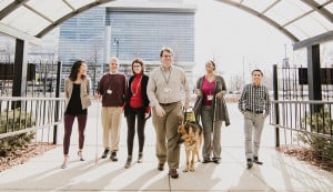 A group of six Chicago Lighthouse employees walk under the glass archway to the building. Two of the employees utilize a white cane and one has a German Shepherd guide dog.