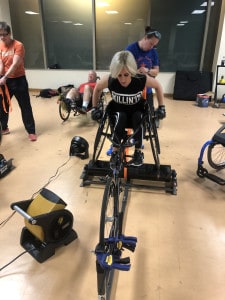 A young woman with disabilities trains for the Chicago Marathon in a gym with others. The woman is using a stationary wheelchair bicycle.