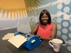 A young woman who is blind is using a Brailler. Behind her is a colorful backdrop with a bold graphic pattern.