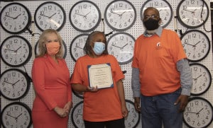 Dr. Janet Szlyk presents a certificate to Alisa Sutton who works as a clock assembler for Chicago Lighthouse Industries. Behind them is a testing wall of clocks.