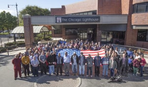 A large group of people (approximately 50) gather around a large USA flag outside of the Chicago Lighthouse. In the front row are employees of the Lighthouse who are Veterans