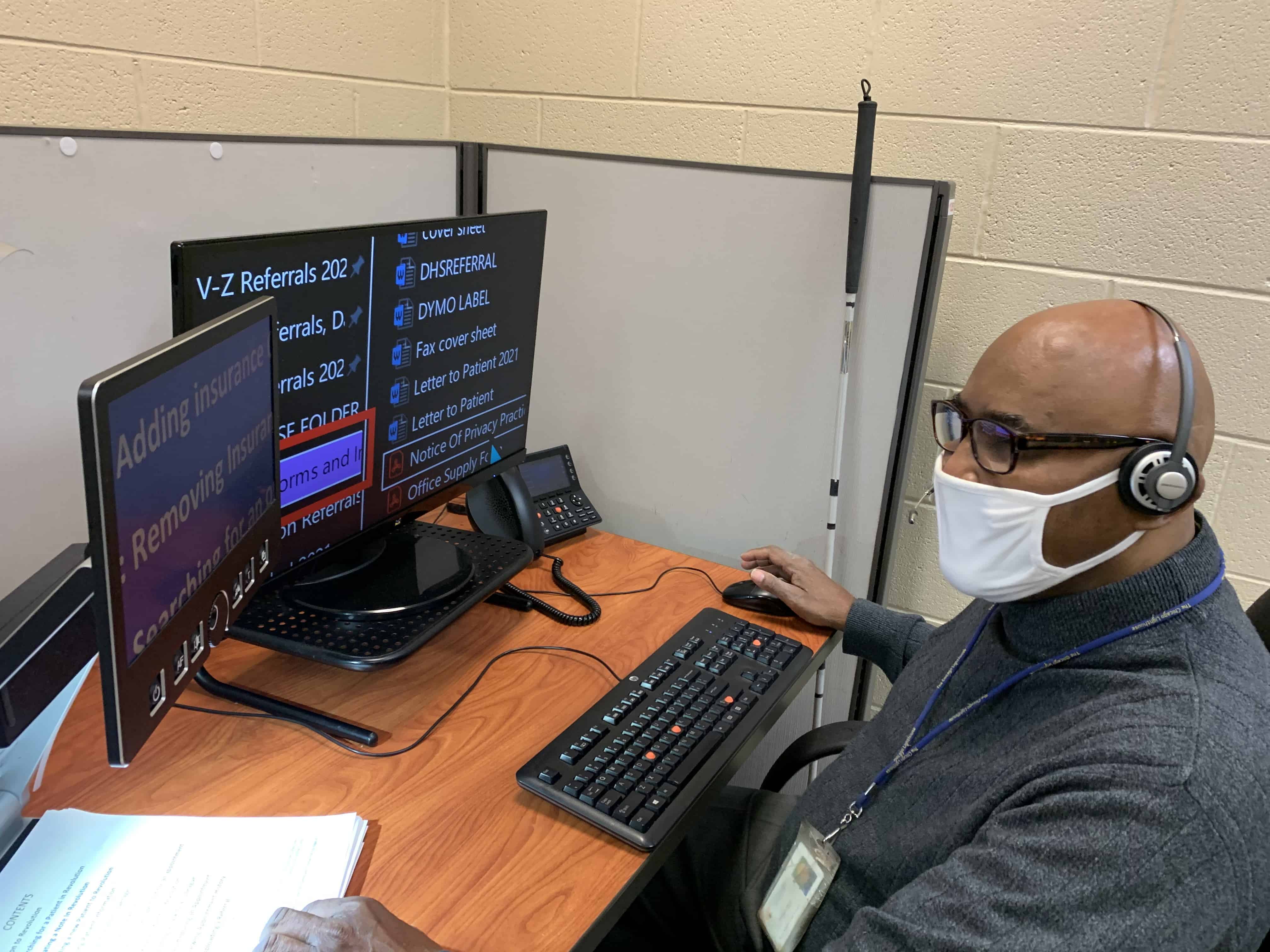 Joseph sitting at a desk in front of his computer, wearing a mask and glasses, with his cane leaning on the desk next to him