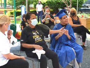 A student of the Childrens Development Center dressed in cap and gown sits with his family before graduation. The family all has t-shirts saying proud (parent, aunt, niece, etc) of a 2021 graduate. All are smiling!