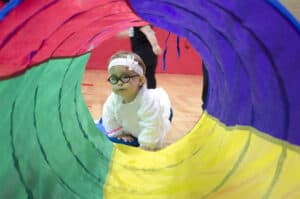 A toddler wearing glasses peeks through a colorful tunnel