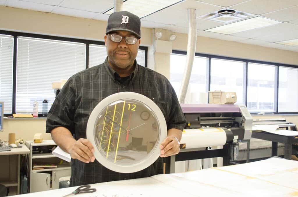 Dave Williams smiles at the camera holding a clock he designed, which has a black background with yellow lines and swirls.