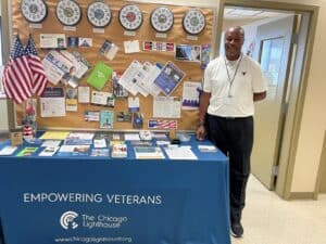 Learnus Ross stands next to a table with flyers on it. It has a tablecloth that says "Empowering Veterans" with the Chicago Lighthouse logo on it. Behind the table is a corkboard with flyers for different Veteran services and six clocks showing the time in different time zones.