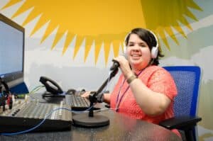 Sandy Murillo sits at a desk with a computer, sound board, and microphone on it. She is wearing  white headphones and is smiling at the camera.