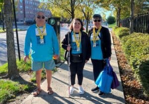 Sandy Murillo and her parents stand on the sidewalk wearing matching blue Rise to Shine Run & Walk shirts and finisher medals. Sandy is holding a white cane.