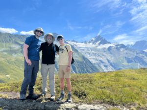 Robert, Andrew, and William Rourke pose for a photo on a beautiful mountaintop. They are wearing hiking outfits and hats and sunglasses.