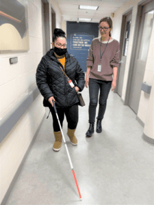 Becca Kelley walks down a hallway next to a client who is practicing using a white cane. 