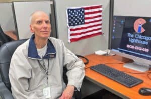 Michael Kanter sits at his desk. An American flag is hanging on his cubicle wall. He is wearing a beige jacket and grey slacks.