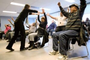 A group of seniors sit in chairs with their arms raised in a chair exercise class. The instructor is adjusting a woman's arms.