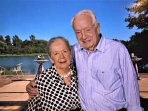 Ann and Walter Nathan standing together outdoors, smiling, with a serene lake and a clear sky in the background. Ann is wearing a polka dot dress and a pearl necklace, and Nathan is dressed in a purple button down shirt.