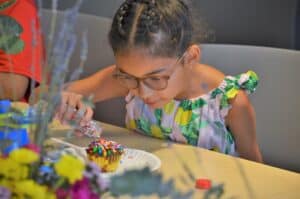 A girl wearing glasses decorates a cupcake with colorful sprinkles at a table filled with crafting supplies.