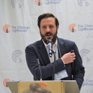 Peter stands behind a podium and speaks. He has a beard and is wearing a navy blazer. 