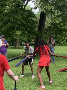 Group of people engaged in a playful foam sword fight outdoors in a park, with several onlookers watching the activity.