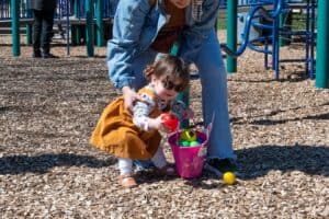 A little girl with pigtails and dark sunglasses in a playground is picking up colorful eggs into a pink basket, assisted by an adult crouching beside her.