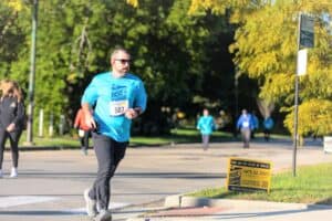 Liam Boyle runs in the 2023 Rise to Shine Run and Walk, wearing a blue 'Rise to Shine' shirt. It is a sunny day, with other participants in the background.