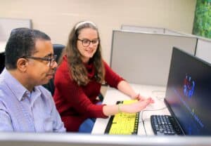 Brittany Davis sits at a desk with a male client in front of a computer screen displaying text "Hi Robert, are you looking for a job right now?" Brittany is gesturing towards the computer screen with a large print yellow keyboard in front of it.