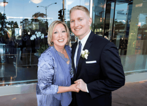 Joy and Joel Bruckman stand together and smile at the camera. They are wearing formalwear, Joy has short blonde hair and is wearing a purple dress. Joel has short blonde hair and is wearing a black suit.