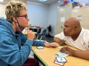 A paraprofessional sits across the table from a CDC student. He is looking at her intently as she holds a finger u p to her face.