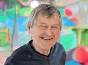 Ed has short gray hair and is wearing a black shirt. He is standing smiling in front of a colorful, abstract art background.