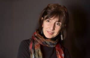 Headshot of Julie Stark. She is wearing a colorful scarf and glasses pushed up on her head, smiling subtly against a dark background.