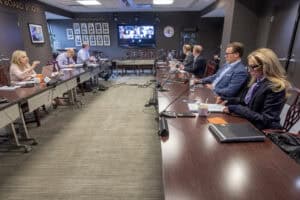 A group of Board of Trustees attend a meeting in a large conference room. Others join virtually on Zoom.