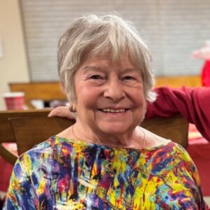Close up shot of Angela, who has short gray hair and a colorful top, smiling warmly.