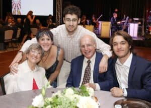 Carol with her family at our Seeing What's Possible Galal. They are seated around a table with white floral decorations with a stage and musicians visible in the background.