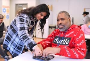 A participant from Adult Living Skills sits at a table in a red 'Hustle' hoodie receives assistance with a Braille clock from Jenny, who is wearing a checked jacket and is learning over the table leaning.